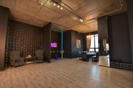 Элегантный лофт с хорошим звуком и балконом. Просторное помещение с высокими потолками и деревянным полом. Черные стены, на одной из которых гирлянда. Зона с креслами и столиком, рядом телевизор. Обеденная зона с большим столом и стульями. Потолочные светильники и люстра. Большие окна с занавесками.