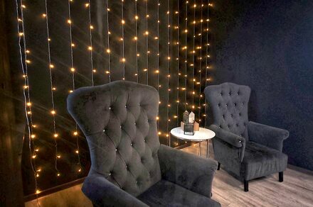 Элегантный лофт с хорошим звуком и балконом. Тёмно-серая комната с двумя мягкими креслами, круглым белым столиком, на котором стоит декоративный светильник. На стене гирлянда из маленьких жёлтых огней. Пол из светлого дерева.