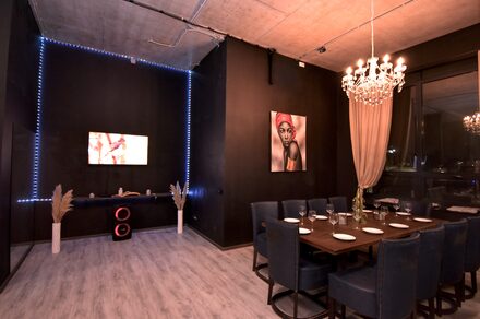 Элегантный лофт с хорошим звуком и балконом. Пространство для мероприятий с деревянным столом и синими стульями, сервированное столовыми приборами и бокалами. Черные стены, подсветка по периметру одной стены, телевизор, картина. Люстра с лампами, светлая штора, декоративные элементы.