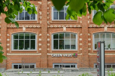 Green Loft(Зеленый Лофт). Фасад кирпичного здания с большими окнами в белой рамке. На уровне первого этажа вывеска "GREEN VKUS CAFE". Перед зданием находится металлическое ограждение и маленькая клумба. На переднем плане видны листья деревьев.