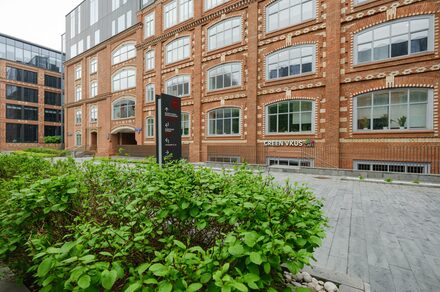 Green Loft(Зеленый Лофт). Красное кирпичное здание с большими окнами. Перед ним тротуар из серых плит и зеленые кусты. У входа вертикальная черная вывеска с текстом. На первом этаже видна надпись «GREEN VKUS».