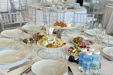 Event  Hall ARCOBALENO (Эвент Холл АРКОБАЛЕНО) . Круглый белоснежный стол, сервированный на банкет. Прозрачные стулья, белая посуда, бокалы, столовые приборы. Центр украшен закусками на металлической подставке. На столе бутылированная вода, овощи и фрукты, кодовые знаки отсутствуют.