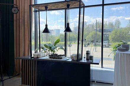 Event  Hall ARCOBALENO (Эвент Холл АРКОБАЛЕНО) . Современный интерьер с барной стойкой и подвесными черными светильниками у панорамного окна, металлическая конструкция с растениями, стеклянные стены, бежевые шторы, белая скатерть на круглом столе с тарелками.