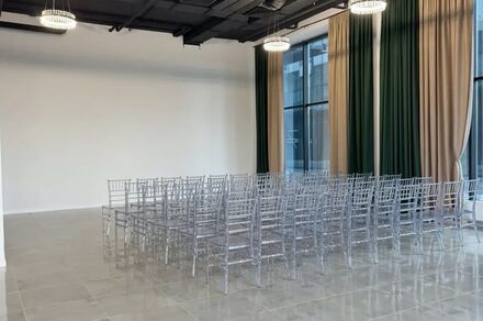 Event  Hall ARCOBALENO (Эвент Холл АРКОБАЛЕНО) . Зал с высокими потолками и большими окнами, занавешенными зелеными и бежевыми шторами. Ряды прозрачных стульев расставлены на сером плиточном полу. Две круглые подвесные лампы на черном потолке. Белая стена без украшений.