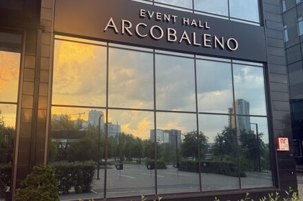 Event  Hall ARCOBALENO (Эвент Холл АРКОБАЛЕНО) . Фасад здания с большими стеклянными окнами, отражающими городской пейзаж с деревьями и небоскребами. Название на фасаде: «EVENT HALL ARCOBALENO». Вывеска «П» на стекле. Цвета: черный, стеклянные поверхности отражают закатное небо.