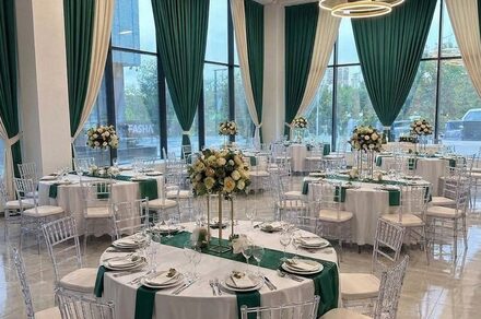 Event  Hall ARCOBALENO (Эвент Холл АРКОБАЛЕНО) . Банкетный зал с круглыми столами, украшенными белыми скатертями и зелеными дорожками. Прозрачные стулья, большие окна с зелеными шторами. Центральные композиции из светлых цветов. Потолочное освещение в виде кольцевых светильников.