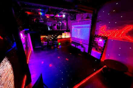 Soul Loft . Тускло освещенное пространство с разноцветными огнями, баром и большим экраном. На стене написано слово Soul в ярко-красных огнях, что создает живую атмосферу, подходящую для встреч или мероприятий. Деревянный пол добавляет теплоты современному декору.