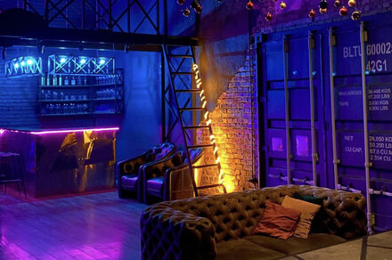 Контраст Loft. Диваны, стол. Декорация в виде дверей большого контейнера. Большая навесная люстра с несколькими лампочками. . Телевизор. Стулья. Свёрнутый экран на стене