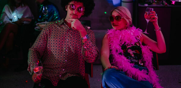 Стилизованный лофт в сине-розовых оттенках. Вечеринка в помещении с диммированным светом. Люди в яркой одежде и аксессуарах сидят с бокалами вина. Задний план освещён разноцветными огнями. Мебель включает стулья и барную стойку. Светильники отсутствуют. Пол тёмный, бетонный или каменный.