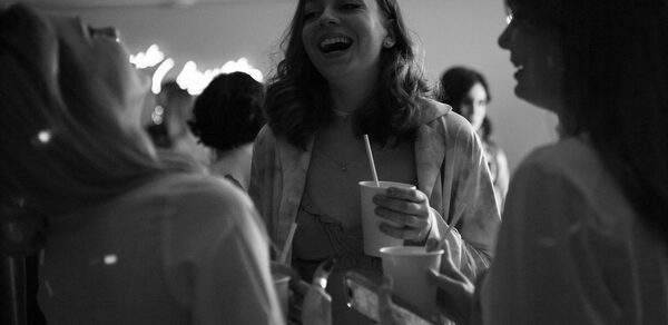 Стилизованный лофт в сине-розовых оттенках. Люди держат напитки в бумажных стаканах. Пространство с приглушенным освещением, в фоне видны светящиеся огни. Черно-белое изображение, лица людей выражают улыбку.