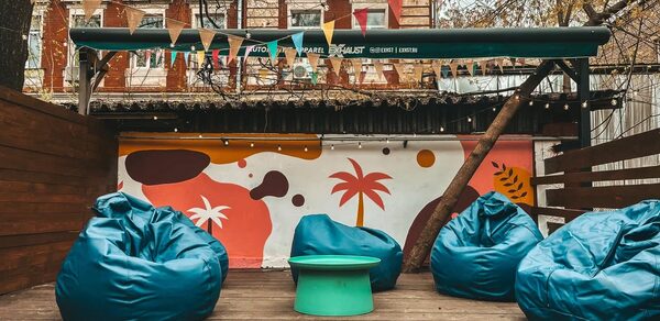 Лофт Surf (Серф). Тропический лофт с большой верандой.. Открытая площадка с деревянным полом, яркие пуфы в виде кресел, круглый стол, разноцветные флажки натянуты сверху, граффити с абстрактными узорами и пальмами на стене, забор из дерева, элементы зелени.