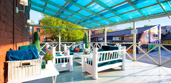 Медный премиальный лофт с летней верандой. Терраса с деревянным полом и деревянной крышей с голубым покрытием. Белая деревянная мебель: диваны и столы. Плетеная корзина и декоративные растения на столе. Стены из красного кирпича, вешалки и фонарики. На заднем плане здания и ограждение из дерева.