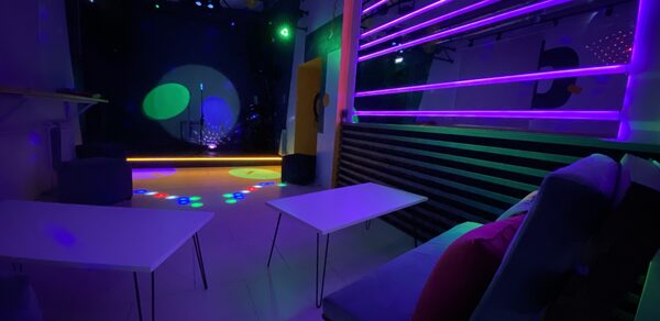 Музыкальный лофт на Тульской. Клубное помещение с фиолетовой и зеленой подсветкой, столами и мягкими креслами, черным стеновым покрытием, сценой с микрофоном и световыми эффектами на полу. На стене вертикальные неоновые полосы, на диванах разноцветные подушки.