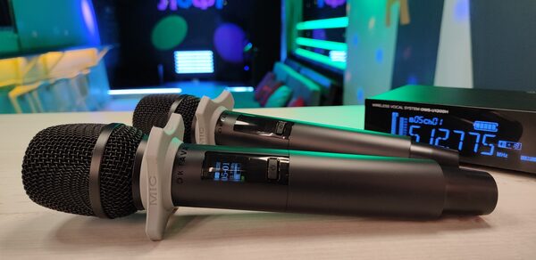 Музыкальный лофт на Тульской. Два беспроводных микрофона на деревянном столе, рядом цифровой приемник с синим дисплеем. На заднем плане разноцветные светодиодные элементы и мягкие диваны, яркие декоративные световые панели на стенах, тусклое разноцветное освещение.