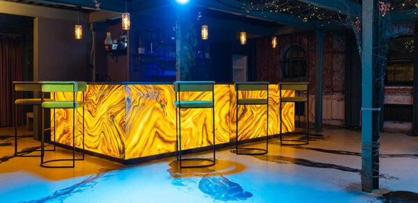 Стилизованный двухэтажный лофт. Барная стойка в индустриальном стиле с подсветкой и мраморным узором. Высокие металлические стулья, свечи на стойке. Потолок с гирляндами, стены из кирпича, пол залит синим светом. Подвесные светильники создают приглушённое освещение.