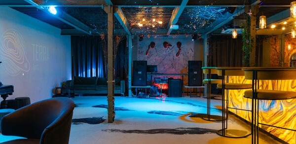 Стилизованный двухэтажный лофт. Креативное многозонное пространство с низкими потолками и открытыми балками. Барная стойка с подсветкой, высокие стулья. Зоны для отдыха с мягкой мебелью. Декоративные потолочные светильники, светодиодное освещение, художественная роспись на стене.