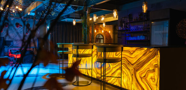 Стилизованный двухэтажный лофт. Современное пространство с барной стойкой из подсвеченного камня, высокими стульями, подвесными светильниками. На потолке гирлянды, деревянные элементы, различные зоны освещения. Интерьер в теплых и холодных тонах, видна декоративная растительность.