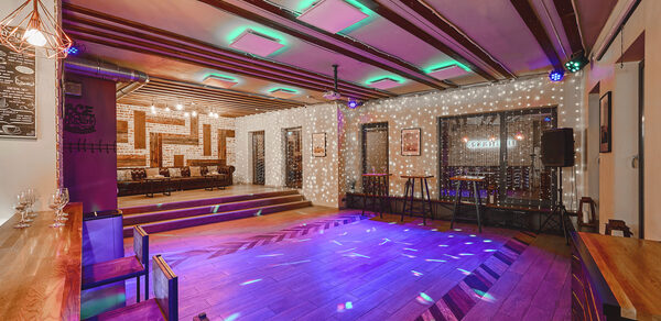 Loft Lodge 6. Просторное помещение с деревянным паркетом и ступеньками на возвышение. Деревянные балки на потолке с подсветкой. Барная стойка с высокими стульями. Неоновое и точечное освещение. Кожаные диваны, стулья, столики и аудиооборудование. Декоративные элементы на стенах.