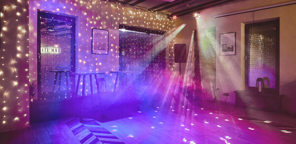 Loft Lodge 6. Праздничное пространство с деревянным полом и мероприятием. Гирлянды свисают со стен, создавая световой эффект. Имеется световое оборудование с разноцветными лучами. На стенах висят картины. Небольшие столы и стулья вдоль стен.