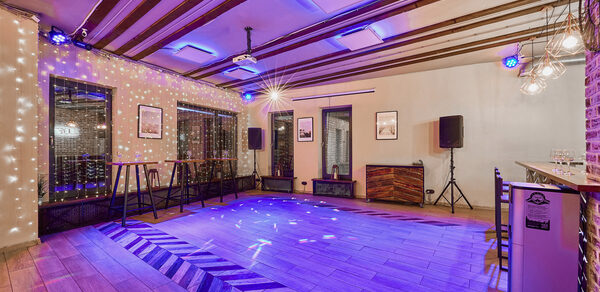 Loft Lodge 6. Просторное помещение с деревянным полом и декоративными балками на потолке. Установлены динамики, проекционное оборудование и светильники. По периметру высокие барные столы и стулья. Настенная отделка из кирпича. На стенах картины в рамках.