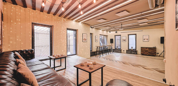 Loft Lodge 6. Просторная комната с деревянным полом и полосатым потолком. Тёмно-коричневый кожаный диван с подушками, два столика. Стены декорированы гирляндами и картинами. Оборудованная барная стойка и аудиосистема. Освещение из подвесных ламп. Трубы на потолке.