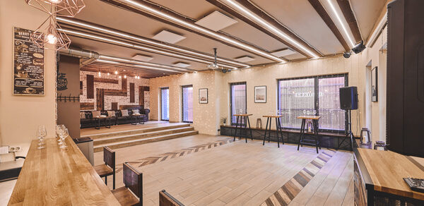 Loft Lodge 6. На фото показано просторное помещение с деревянным полом и ступенчатым подиумом. В центре сцена с диванами. Оборудование: нижние столы, стулья, барная стойка, динамик. Оформление: кирпичная стена, потолочные светильники. Цвета теплые, состояние хорошее.