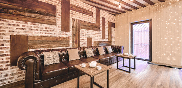 Loft Lodge 6. Комната с кирпичными стенами и деревянными вставками, деревянный пол. Большой кожаный диван коричневого цвета с подушками, перед ним два столика с посудой. Освещение — лампочки на проводах, гирлянда на стене. Цвета тёплые, интерьер в отличном состоянии.