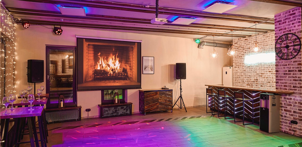 Loft Lodge 6. Это просторное помещение с деревянным полом и декором из кирпича. На стене изображение камина. Имеется барная стойка, столы и стулья. Освещение состоит из ламп и цветных прожекторов. Присутствуют аудиоколонки. На стене часы. Состояние ухоженное.