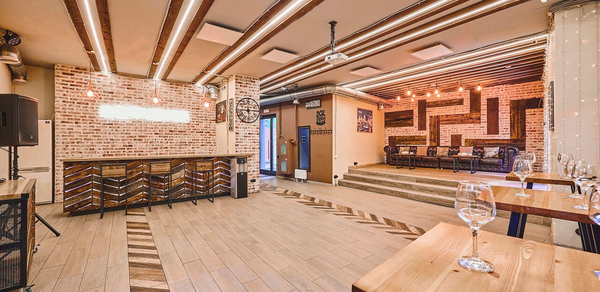 Loft Lodge 6. Это интерьер кафе или ресторана с кирпичными стенами. В центре деревянная барная стойка с высокими стульями. Потолок украшен деревянными балками и лампами. На полу плитка с узором. Присутствуют деревянные столы с бокалами. Общее освещение теплое.