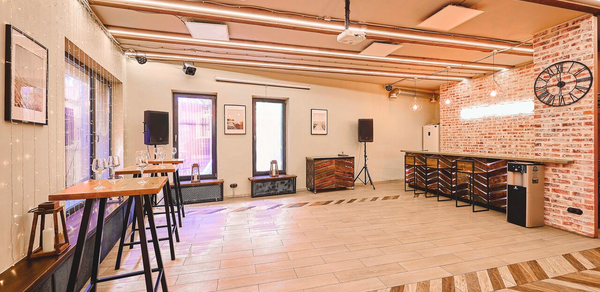 Loft Lodge 6. Пространство представляет собой зал с высоким потолком и плиточным полом. Стены оформлены под кирпич. Есть высокие деревянные столы и черные стулья. На стенах картины и часы. Освещение создано с помощью подвесных ламп и неоновой вывески.