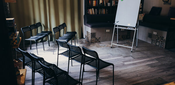 Лофт с возможностью зонирования. Небольшое помещение с деревянным полом, черные стулья в два ряда, флипчарт на треноге. В углу книжные полки и диваны с подушками. Тяжелые шторы на окне. Стены светло-серые, с декоративными вставками. Освещение мягкое.