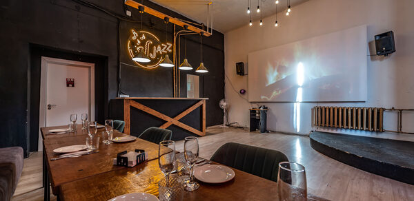 Rubin Loft (Рубин Лофт). Зал Джаз. Ресторанное пространство с длинным деревянным столом, сервированным тарелками и бокалами. На стене вывеска и проектор с изображением. Освещение от ламп накаливания. Черные стены, пол светлый, есть сцена.
