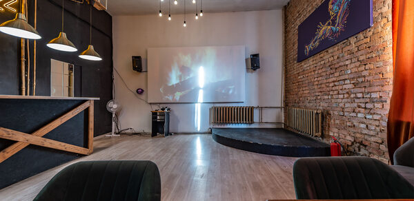 Rubin Loft (Рубин Лофт). Зал Джаз. Лофтовое помещение с кирпичной стеной, проектор на стене, сцена, стулья, украшенные текстилем; две люстры, барная стойка с деревянной отделкой и три подвесных светильника. Пол светлый, справа красные шторы.