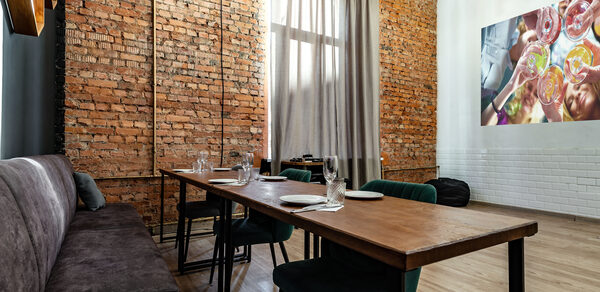 Rubin Loft (Рубин Лофт). Лофт средний. Лофт-пространство с кирпичными и белыми стенами, деревянный стол, стулья с тёмной обивкой, длинный мягкий диван, подвесное освещение, большая фотография на стене, высокий потолок, деревянный пол, окно с занавесками.