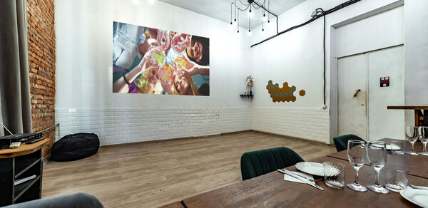 Rubin Loft (Рубин Лофт). Лофт средний. Просторная комната с кирпичной и белой плиточной стенами. В центре проекция на стене. Подвесные светильники на черных проводах. На переднем плане деревянный стол с сервировкой, стулья, бокалы, тарелки, приборы. Деревянный пол, пуфик.