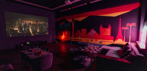 Зал Лас-вегас. Лофт-зал в американском стиле . Темное помещение с мягкой мебелью и проектором, проецирующим изображение города на стене. Яркий граффити-силуэт городского горизонта на стене. Низкие столики с посудой. Настенное освещение создает уютную атмосферу.