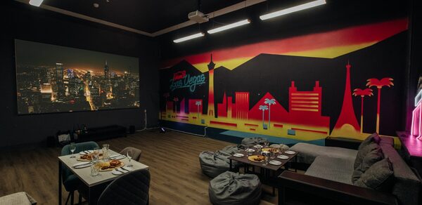Зал Лас-вегас. Лофт-зал в американском стиле . Темная комната с проектором, на стене изображение города. Стена украшена ярким рисунком городского пейзажа с пальмами. В центре стоят стол и стулья с готовыми блюдами. Справа диваны и кресла-мешки. Освещение подвесное, пол светлый.
