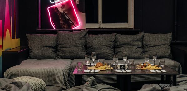 Rubin Loft (Рубин Лофт). Зал Лас-вегас. Большая комната с темными стенами, серые диваны с подушками, низкий стол с тарелками и стаканами. Неоновая вывеска в форме снимка человека, светлое окно, несколько кресел, мягкое освещение.