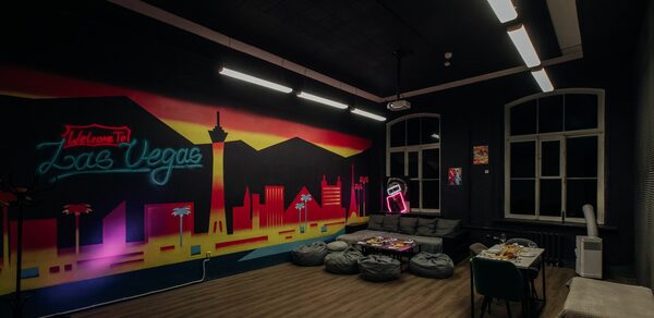 Зал Лас-вегас. Лофт-зал в американском стиле . Темное помещение с граффити на стене, изображающем ночной Лас-Вегас. Пол деревянный, потолочные светильники продолговатые. В углу зона с серыми пуфами и столиком. Под неоновым знаком стоят кушетка и стул. Большие окна без штор.