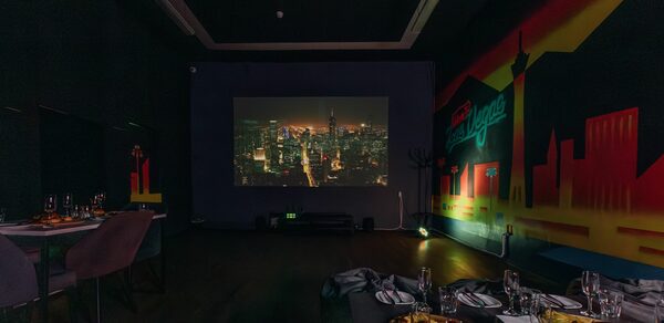 Rubin Loft (Рубин Лофт). Зал Лас-вегас. Тёмная комната с проектором на стене, показывающим городской пейзаж. На стенах граффити со зданиями. Несколько столов с посудой и сервированными блюдами. Мягкие стулья серого цвета, тёмный деревянный пол. Слабое освещение.