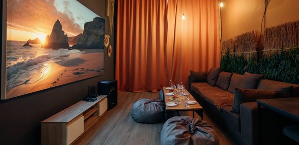 Rubin Loft (Рубин Лофт). Зал Ямайка. Кейтеринг-зона с деревянным полом, оранжевыми шторами, коричневым диваном, двумя креслами-грушами, низким столом с сервировкой. На стене большой пейзажный экран. На потолке лампы в подвесных светильниках, настенное растение.