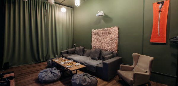 Rubin Loft (Рубин Лофт). Зал Китай. Зал с зелеными стенами и шторками, деревянный пол. Серый диван, кресло, два мягких пуфа, журнальный столик с посудой. Подвесные светильники, декоративная панель на стене. Красный постер справа, проектор на потолке.