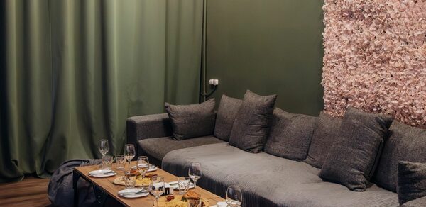 Rubin Loft (Рубин Лофт). Зал Китай. Просторная гостиная с серым угловым диваном и низким деревянным столом, сервированным бокалами и закусками. Зелёные шторы, стена с покрытием из розовых цветов. Деревянный пол. Мягкое освещение. Комната оформлена в спокойных тонах.