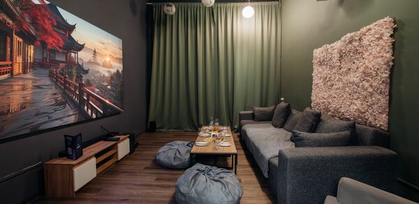 Rubin Loft (Рубин Лофт). Зал Китай. Компактное помещение с современным интерьером. Мягкий серый диван, пуфы, деревянный стол. Большой экран на стене, ковровое панно. Потолочные светильники, зелёные шторы. Напольное покрытие из дерева. Белый проектор на потолке.