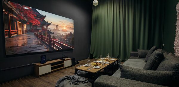 Rubin Loft (Рубин Лофт). Зал Китай. Кинотеатр с серым диваном и пуфом, деревянный пол, на стене большой экран с изображением пейзажа. Зеленые шторы на окне, на потолке подвесные светильники. Перед диваном низкий столик с сервировкой бокалами и тарелками.
