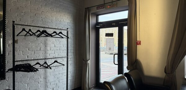 Лофт Повод Есть. Гардеробная зона с белой кирпичной стеной и металлической вешалкой с черными плечиками. Справа стеклянная дверь с вывесками «вход» и «выход», занавески на окнах. Деревянный пол, кожаные кресла темного цвета. Светильников не видно.