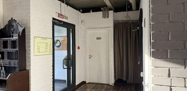 Лофт Повод Есть. Коридор с белыми кирпичными стенами, деревянным полом, стеклянной дверью с черной рамой, рядом вешалка с коричневыми занавесками. На стене желтая схема. Потолок с открытыми коммуникациями и висящими лампами.