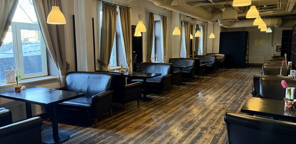 Лофт Повод Есть. Просторное помещение с деревянным полом, длинный ряд черных кожаных диванов и столиков, окна с занавесками, подвесные светильники с теплым светом, белые стены, вентиляционные трубы на потолке, цветы на столах.