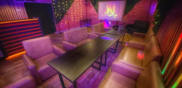 Forest(Лес). Каминный зал с кирпичными стенами и деревянным полом. Диваны коричневого цвета расставлены вокруг черного прямоугольного стола. На стене экран с изображением камина. Светодиодная подсветка на стенах и барной стойке. Потолочные светильники.