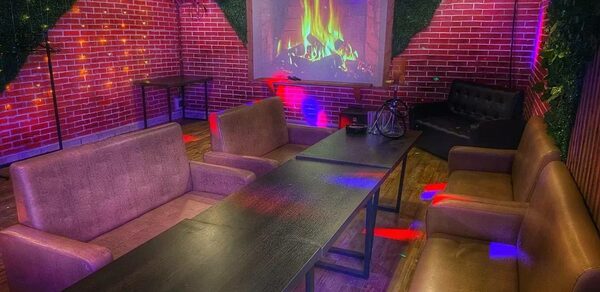 Forest(Лес). Уютное помещение с кирпичными стенами, темными деревянными столами и мягкими коричневыми диванами. На стене проекция камина. Пол из дерева. Разноцветное освещение создает атмосферу. В углу черный диван. На стенах зелень.